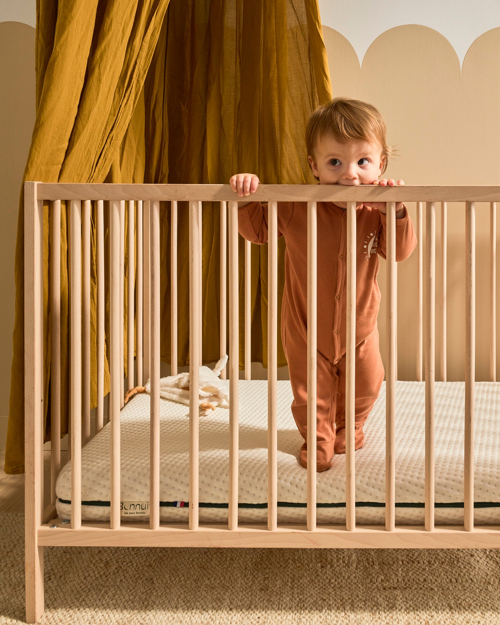 Bébé s’appuyant sur le rebord de son lit à barreaux, posé sur un matelas Bonnuit, les mains accrochées aux barreaux