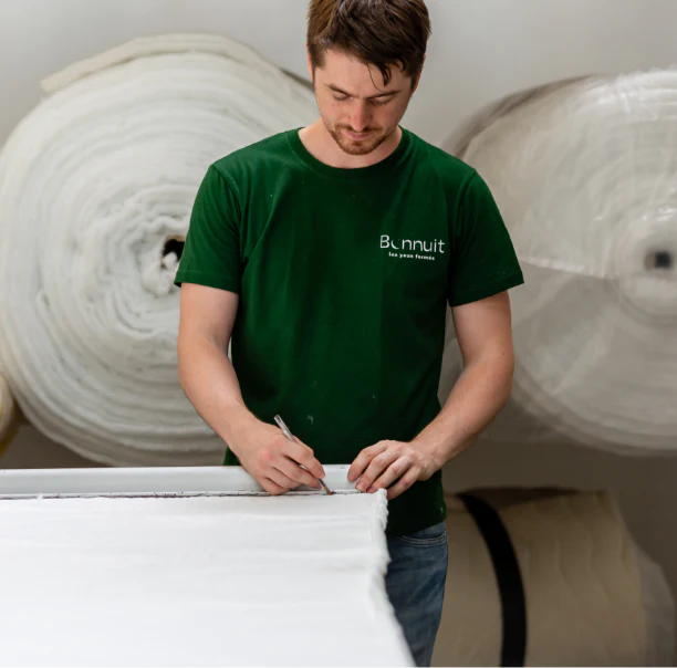 Couturier Bonnuit en train de tracer sur du tissu avant découpe, vêtu d’un t-shirt vert, avec de gros rouleaux en arrière-plan
