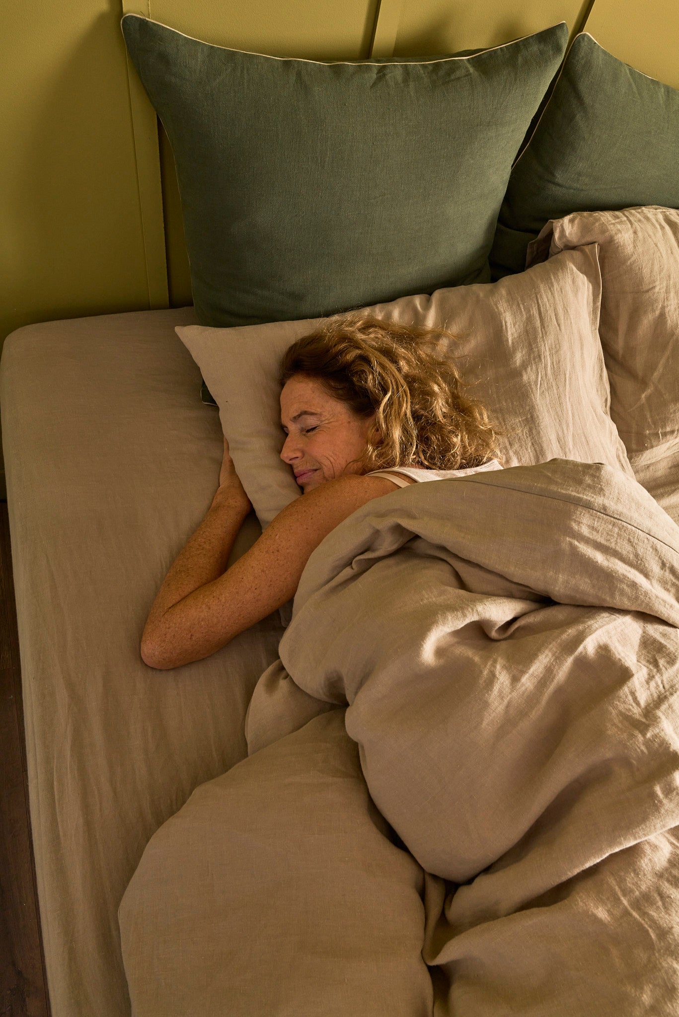 Femme apaisée allongée dans un lit Bonnuit avec des oreillers en évidence et du linge de lit en lin lavé beige