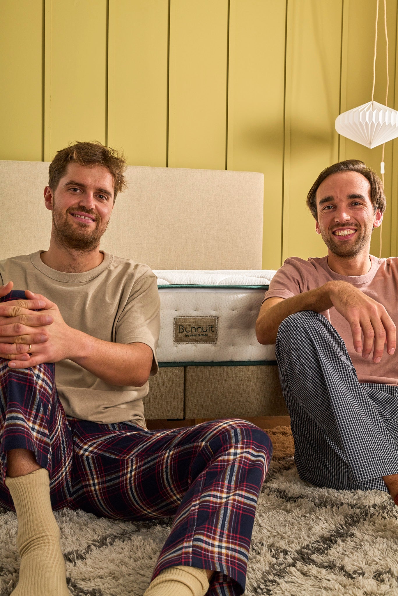 Les fondateurs de Bonnuit assis en pyjama devant un lit de la marque, symbolisant simplicité, engagement et proximité