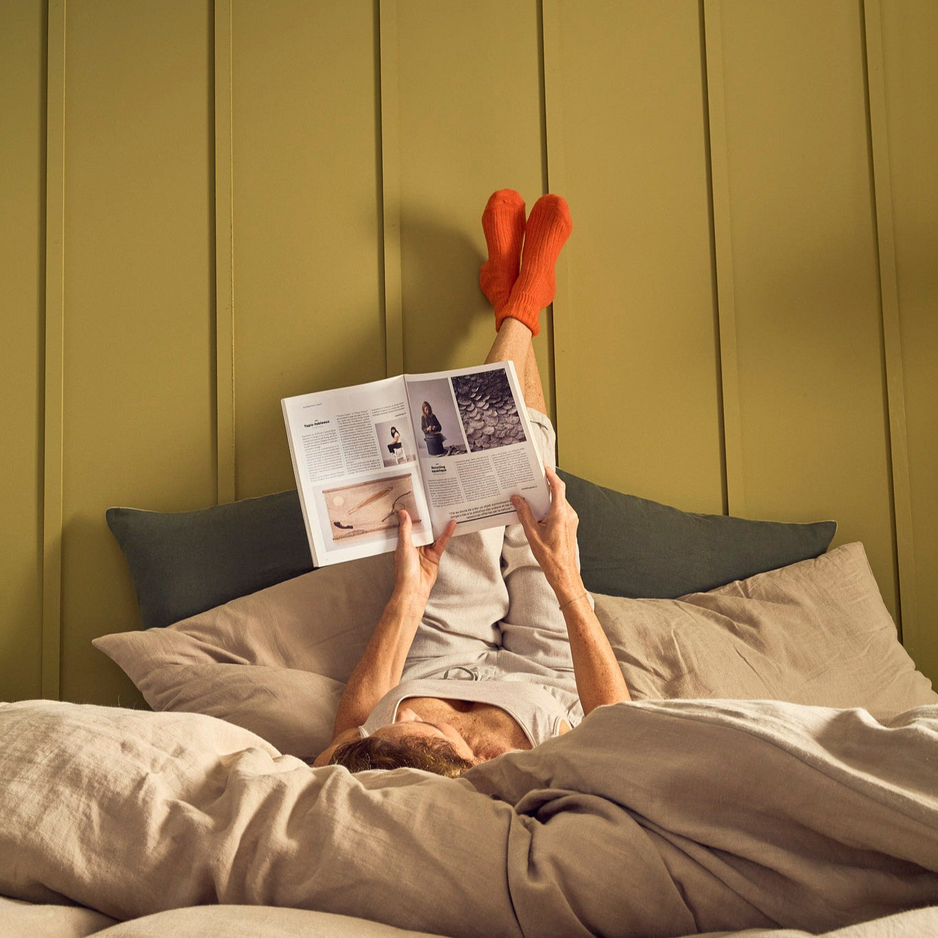 Personne lisant un livre sur le matelas Le Naturel, parure en lin lavé beige, pieds posés contre le mur
