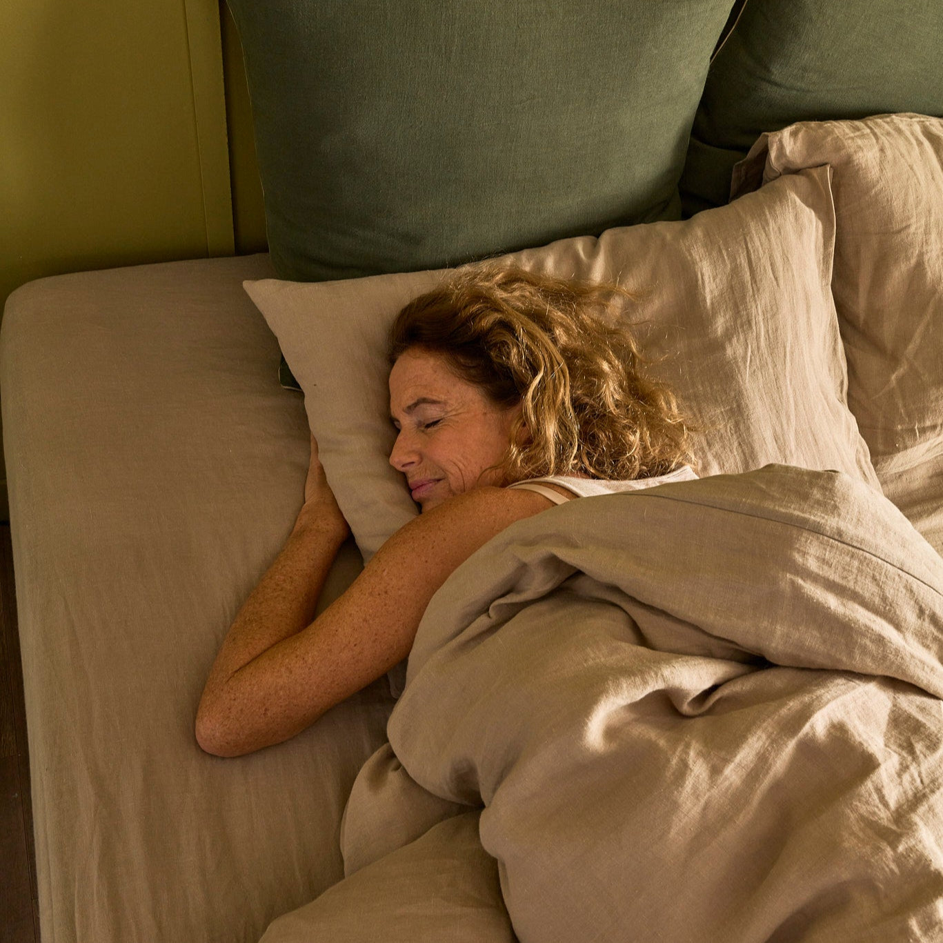 Femme allongée sur le ventre sur le matelas Le Nuage Bonnuit, blottie contre son oreiller, parure en lin lavé beige