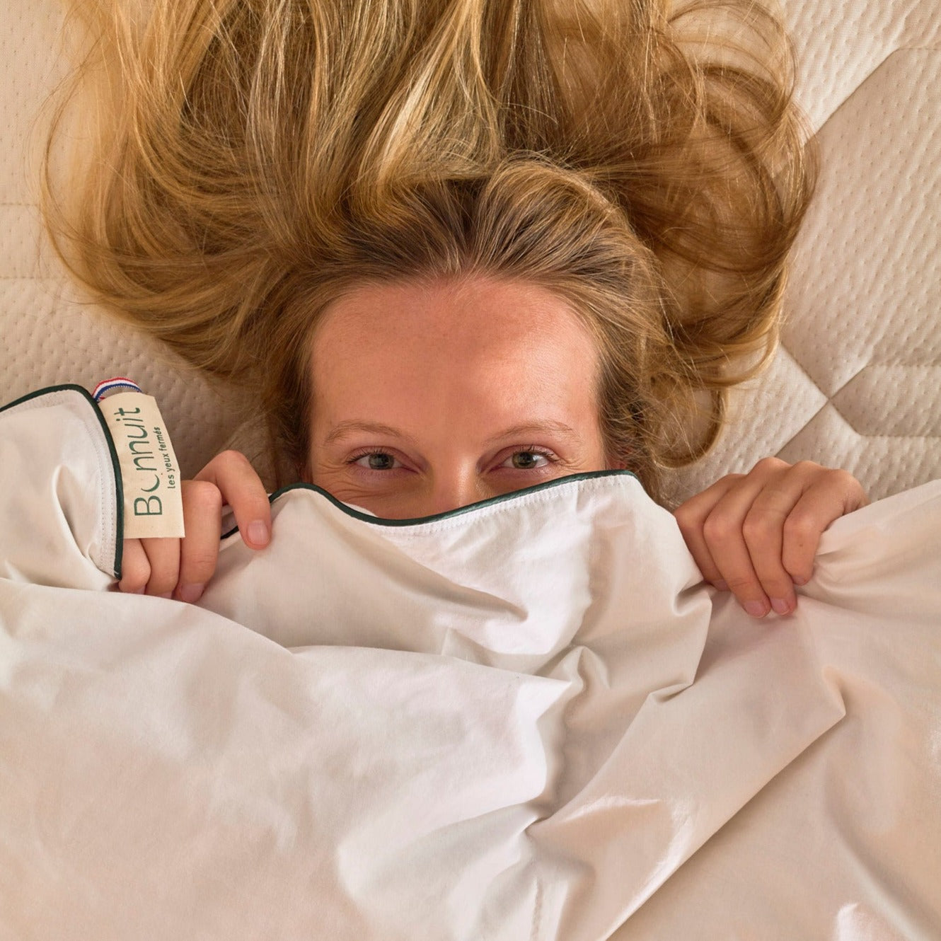 Femme blottie sous la couette Polyvalente remontée jusqu’aux yeux, étiquette Bonnuit et drapeau BBR visibles