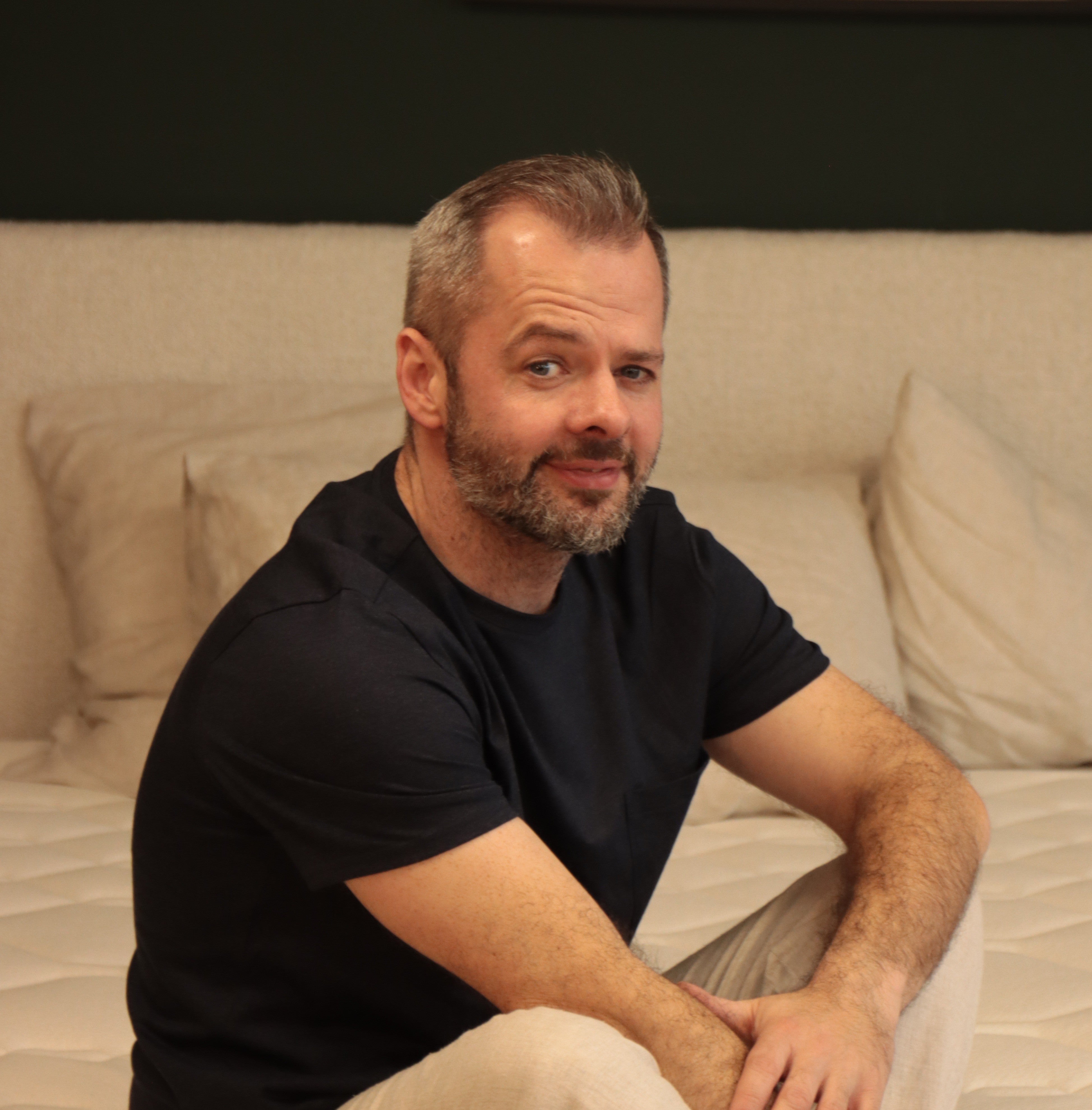 David, tapissier Bonnuit, assis en pyjama sur un lit, avec un sourire tranquille et les mains posées sur les genoux