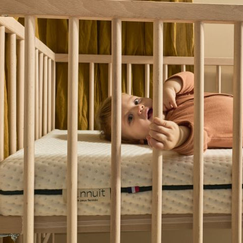 Bébé allongé dans son lit à barreaux avec le matelas Mini Mousse, main sur le barreau, housse claire visible