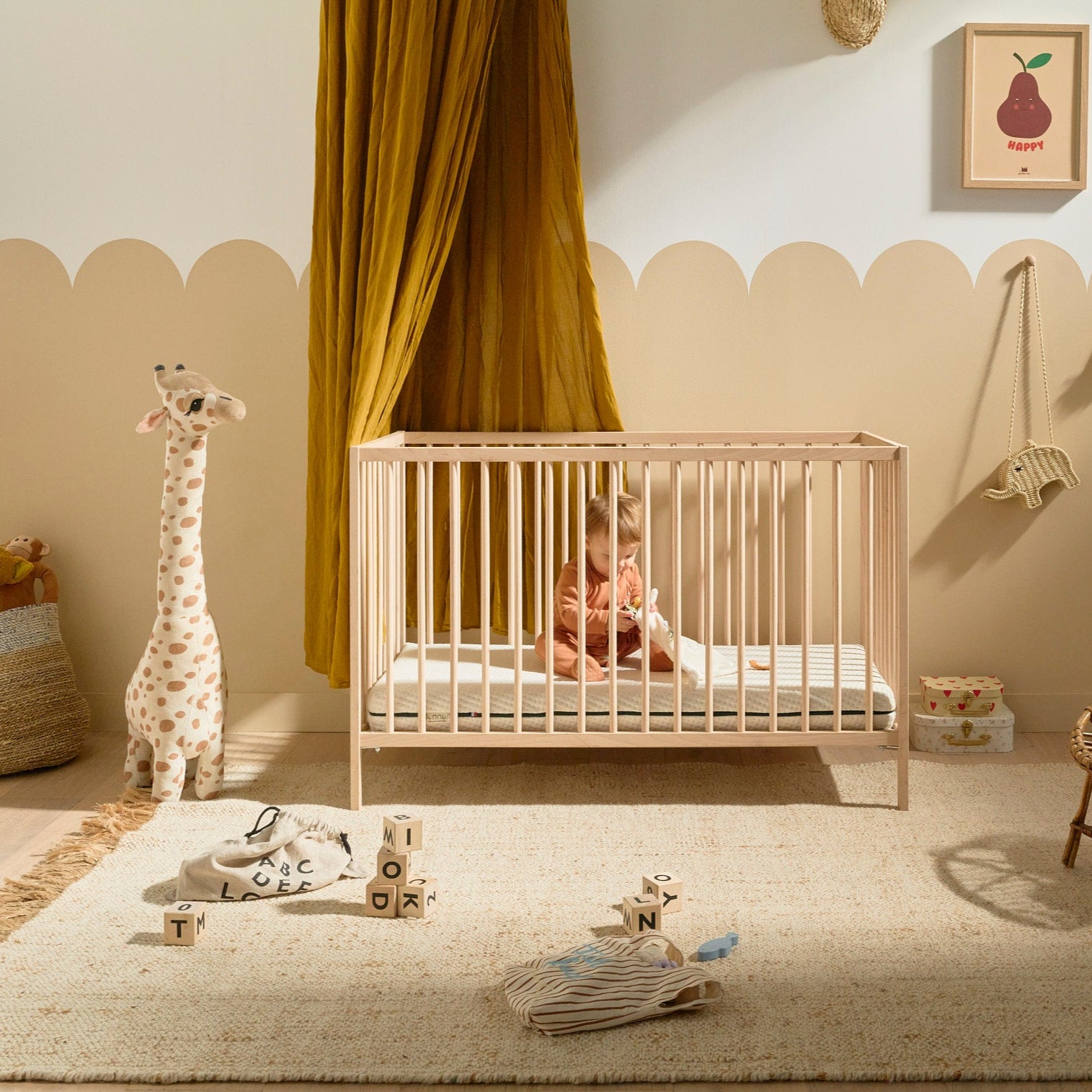 Bébé assis dans son lit à barreaux avec le matelas Mini Mousse, déco cosy de la chambre en arrière-plan