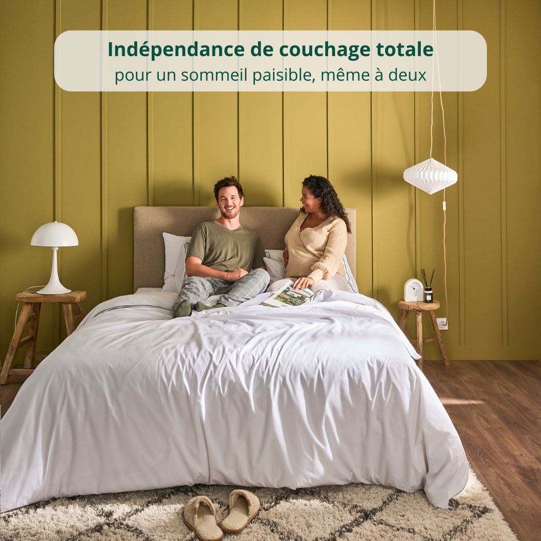 Un couple assis sur un matelas Original Bonnuit habillé d’une parure blanche, adossé au mur avec bannière illustrant l'indépendance de couchage