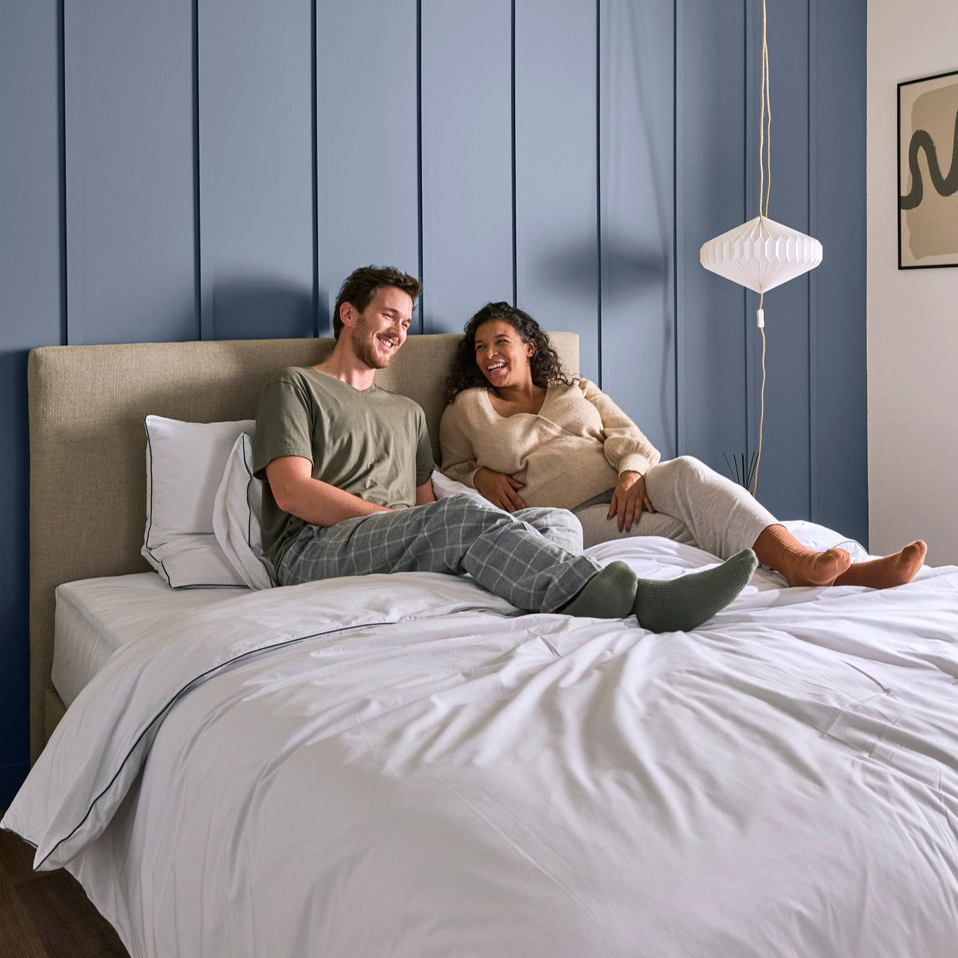Couple adossé à la tête de lit sur la literie Bonnuit, linge de lit blanc en percale avec liseré vert, ambiance cosy