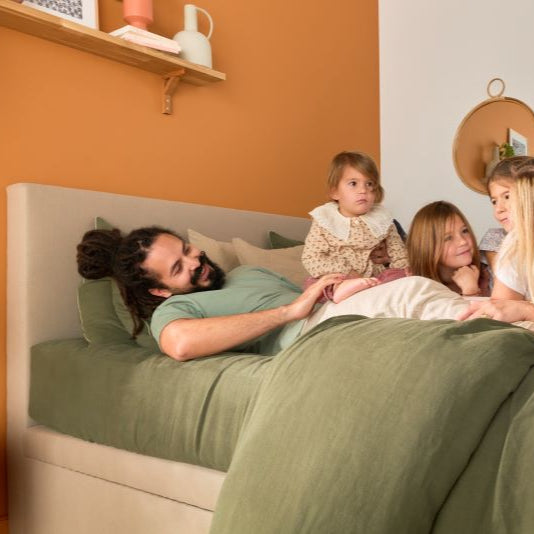 Famille qui s’amuse sur le lit Bonnuit, tête de lit Impériale à l’arrière, linge de lit en lin lavé vert