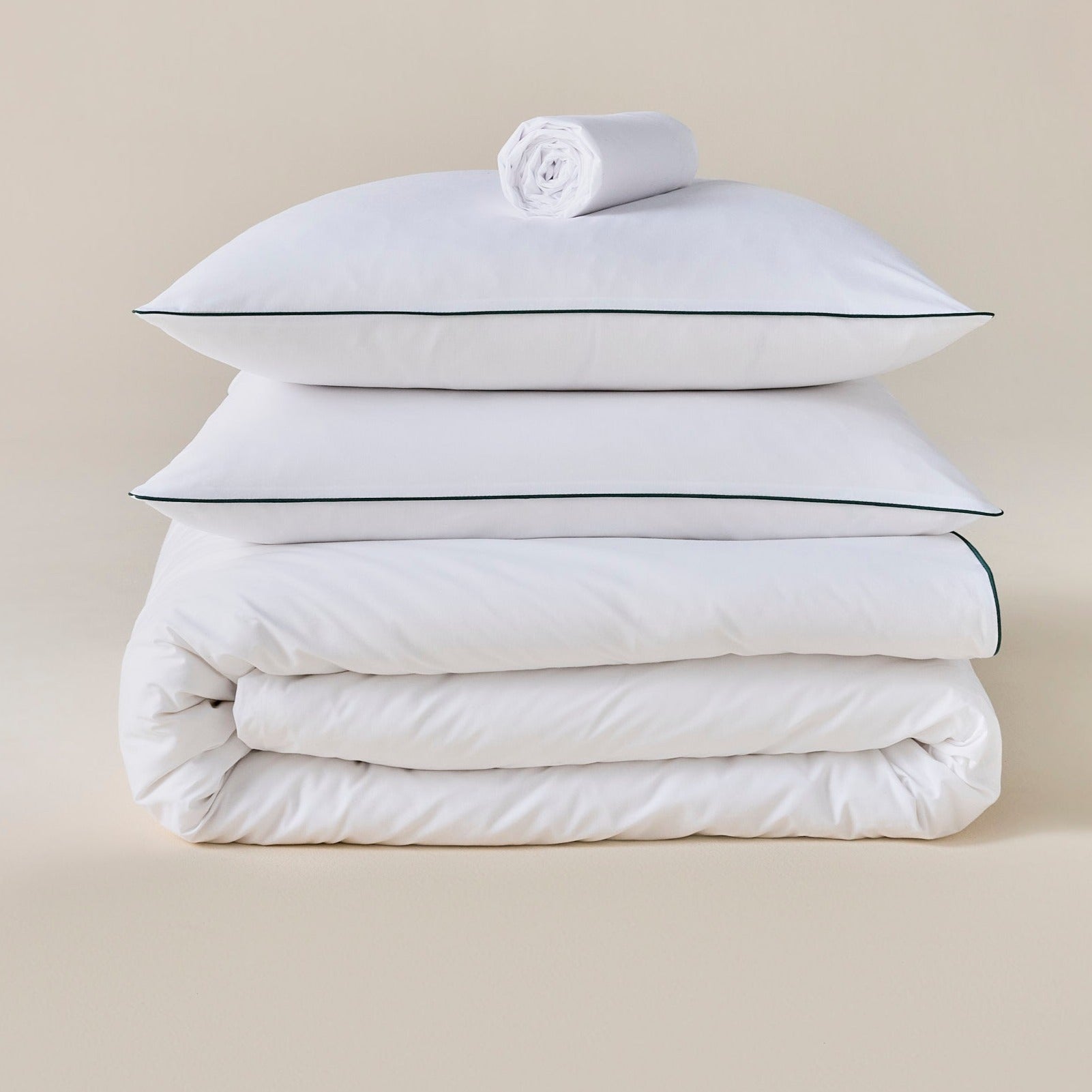 Parure de lit en percale blanche avec liseré vert, oreillers sur la couette et drap roulé posé au-dessus, fond neutre