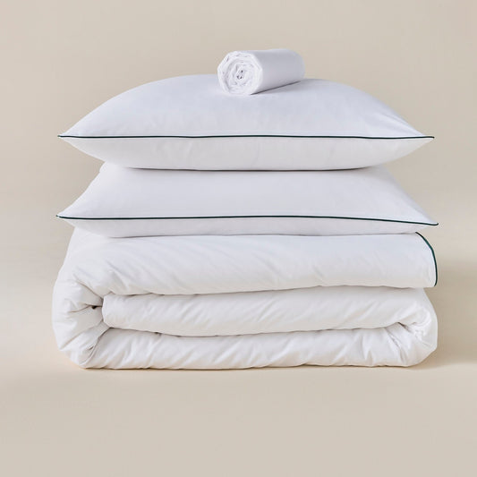 Parure de lit en percale blanche avec liseré vert, oreillers sur la couette et drap roulé posé au-dessus, fond neutre