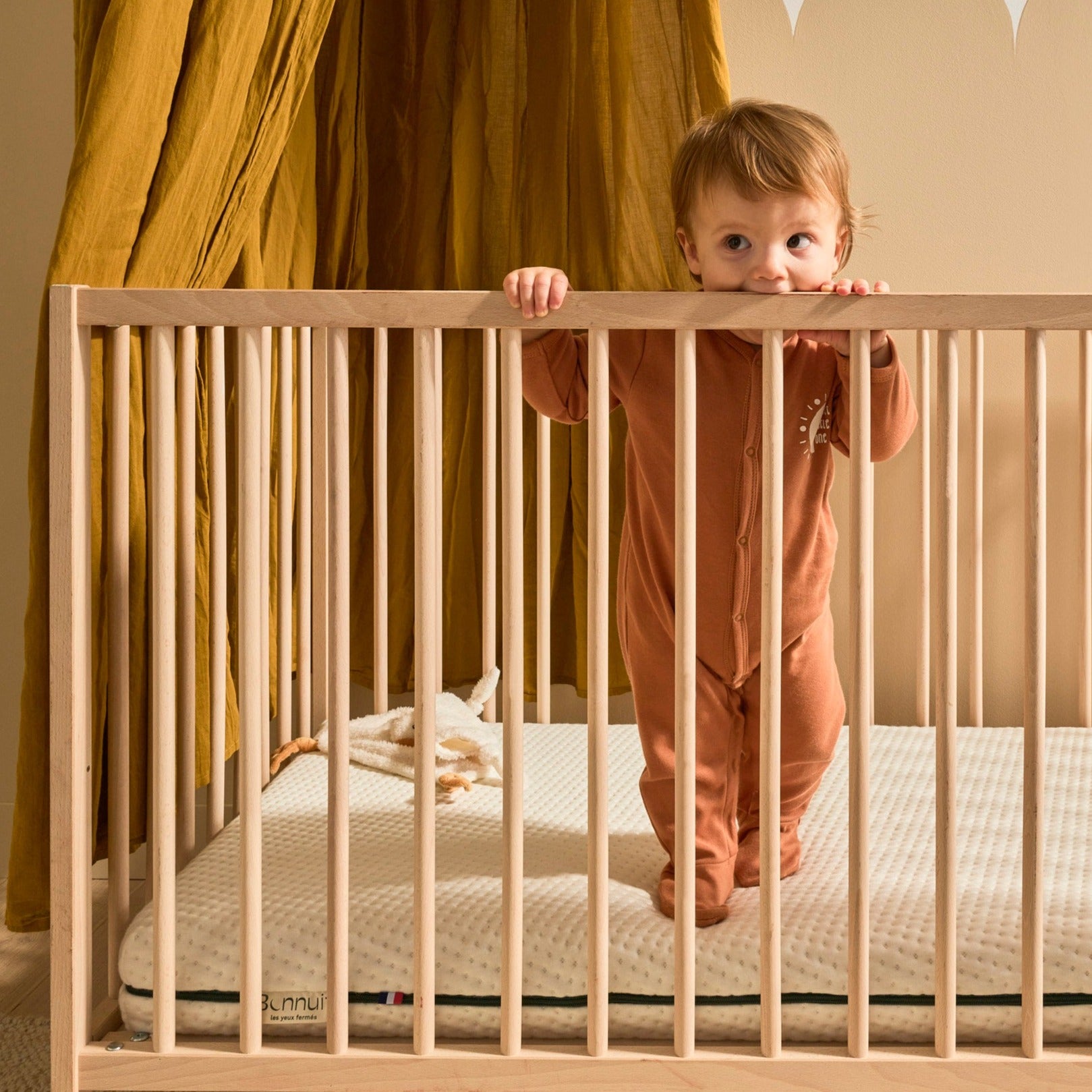 Bébé debout dans son lit à barreaux avec le matelas Évolutif Bonnuit, housse claire et étiquette visible